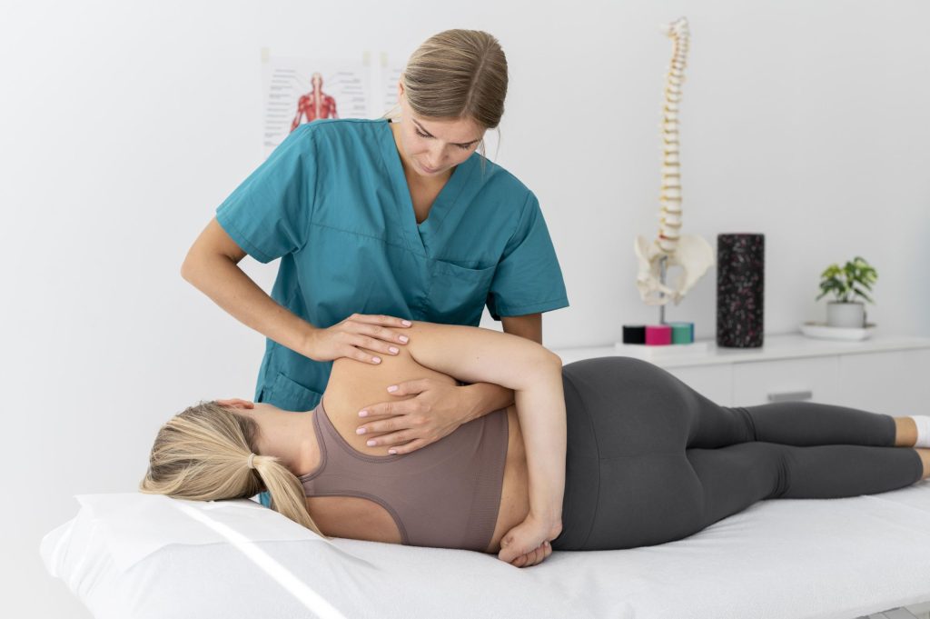 A chiropractor in a teal uniform performing a spinal adjustment on a woman lying on her side in a clinical setting, with anatomical models visible in the background.