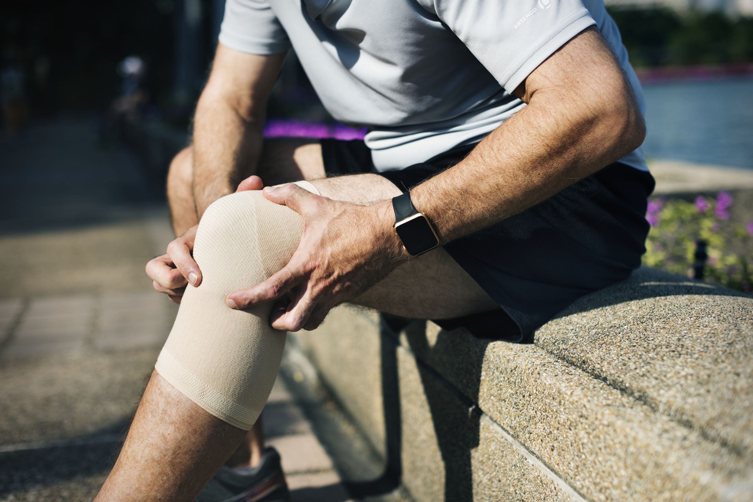 A man in sportswear sits on a stone ledge outdoors, clutching his knee which is supported by a compression sleeve, possibly due to an injury or discomfort after physical activity.