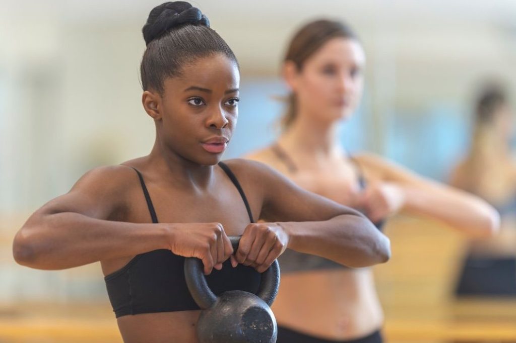 Two women in athletic wear lifting kettlebells during a strength training workout in a fitness studio, with the woman in the foreground appearing focused and determined.