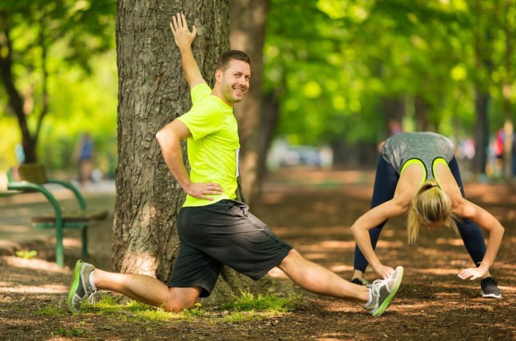 A man and woman stretching in a park; the man is doing a side lunge while using a tree for support, and the woman is bending forward to stretch her hamstrings.