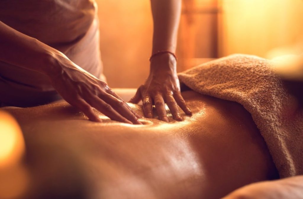 A person receiving a back massage in a dimly lit, serene setting, with hands applying oil and gentle pressure.