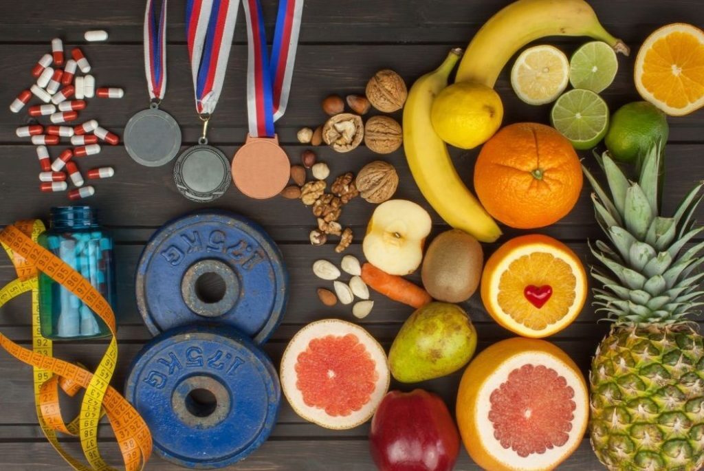 Flat lay of healthy fruits, nuts, and vegetables next to fitness items like dumbbells, a water bottle, a measuring tape, medals, and capsules, symbolising health, nutrition, and sports performance.