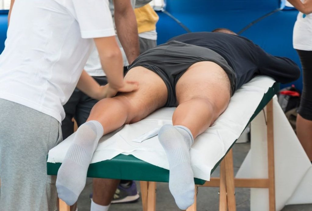 A person lying face down on a massage table receiving a leg massage from a therapist, likely as part of a sports recovery routine at an event or race.