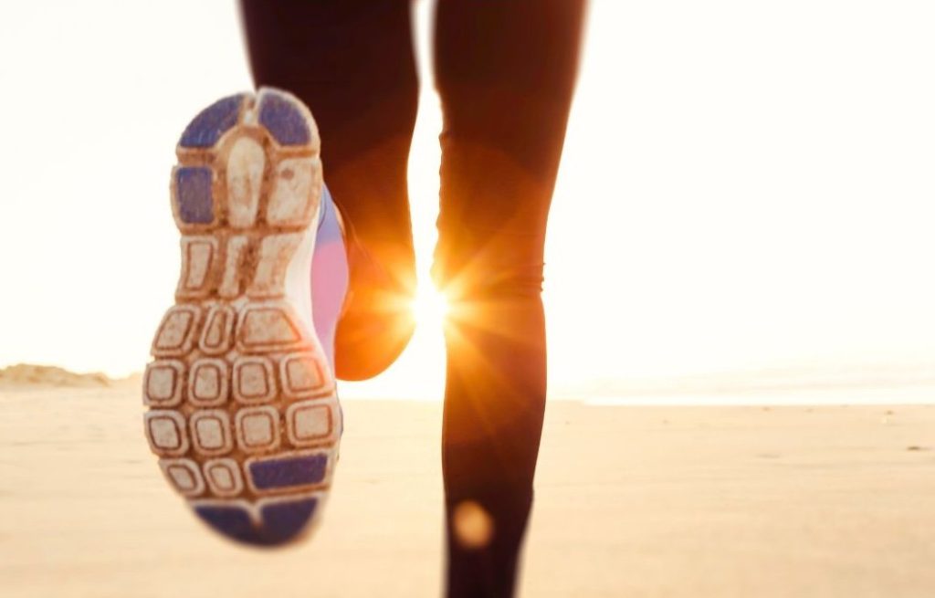 Close-up of a runner's legs and shoe sole as they jog along a sandy beach at sunrise, with sunlight glowing between their legs.