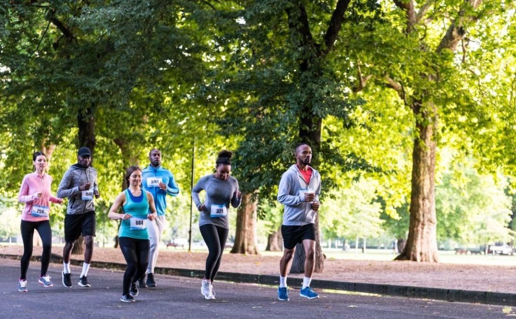 A diverse group of people running together through a park, wearing numbered bibs, likely participating in a community race or fun run event.