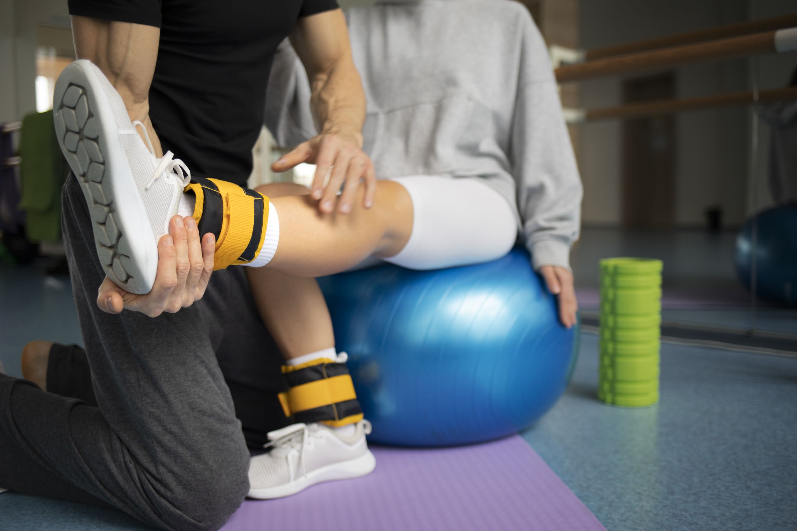 A physiotherapist assisting a patient with a leg exercise using ankle weights and a stability ball in a rehabilitation setting.