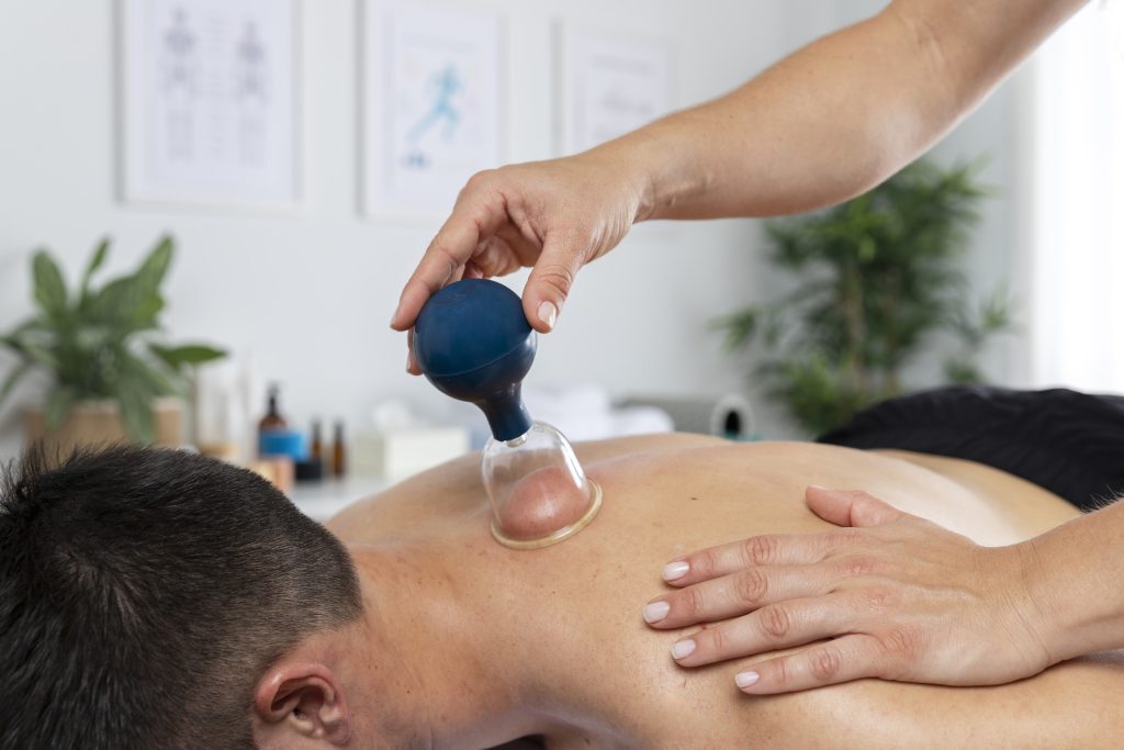 A therapist uses a cupping device on a patient's back, applying suction therapy in a relaxing treatment room. The patient lies face down, receiving gentle care.