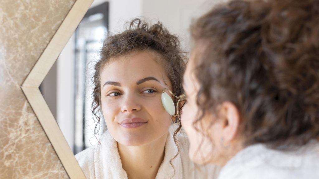 Woman using a jade roller on her cheek while looking into a mirror.