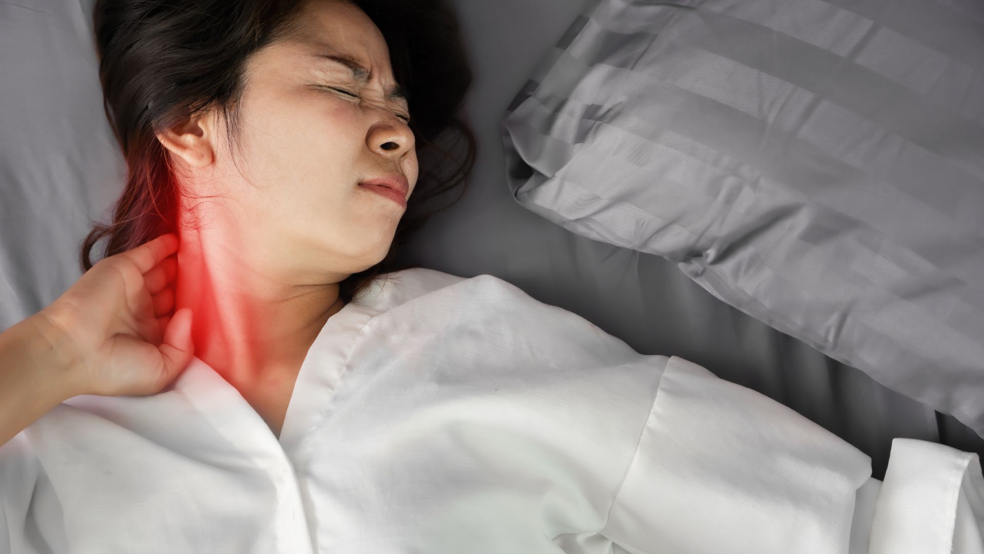 A woman lying in bed wearing a white shirt, grimacing in pain while holding her neck, with a red glow indicating discomfort.