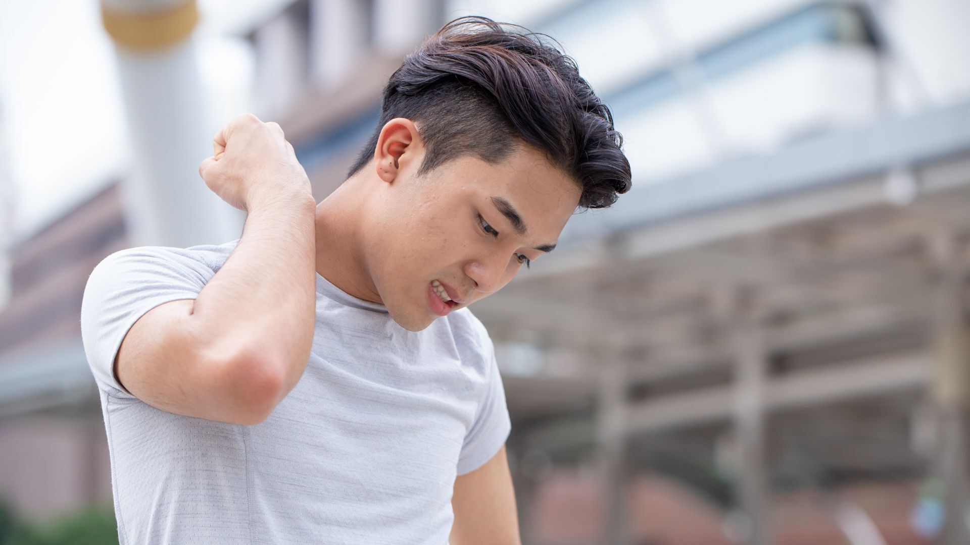 A young man in a white t-shirt outdoors, holding his shoulder with a pained expression.