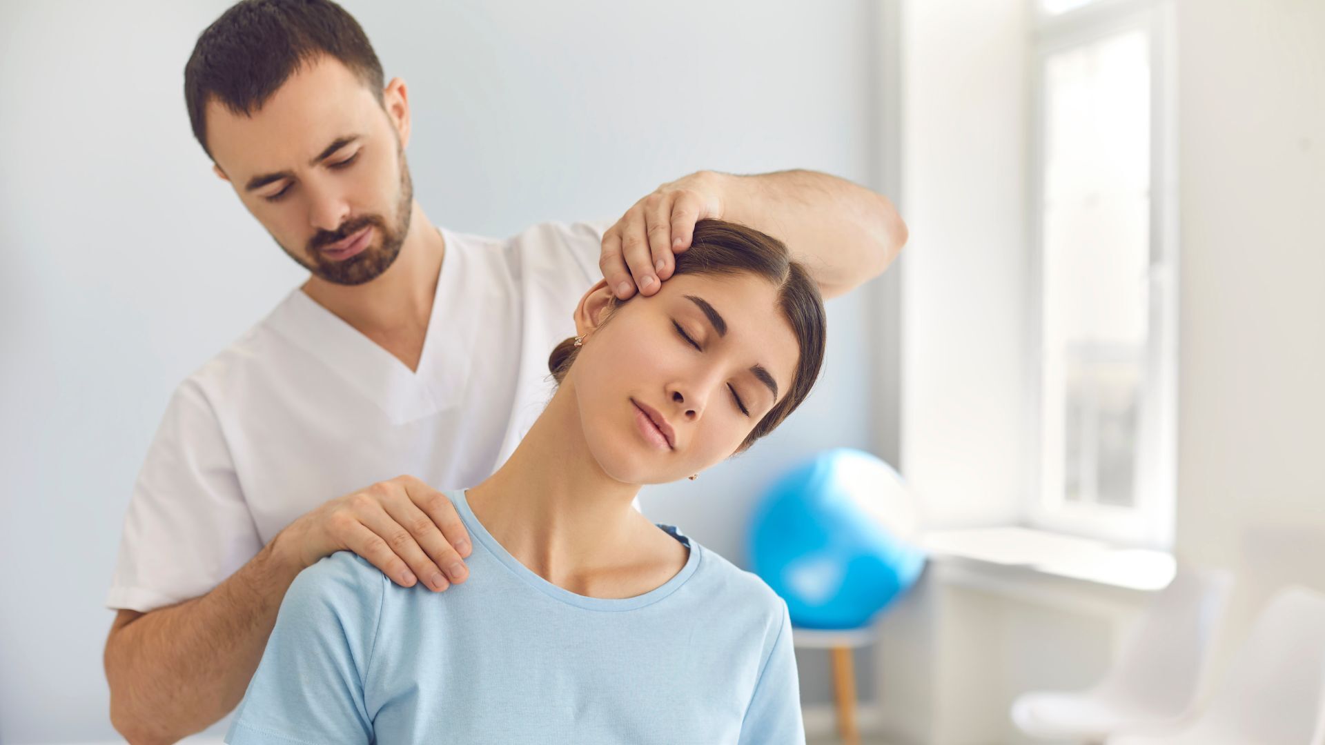 A physiotherapist in a white uniform performing a neck adjustment on a relaxed female patient wearing a light blue shirt in a bright clinic.