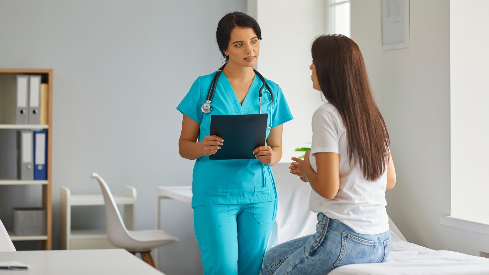 A female healthcare professional in blue scrubs talking to a patient in a clinical setting.