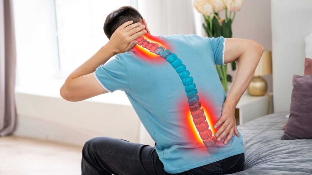 A man sitting on a bed holding his neck and lower back, with a digitally highlighted spine indicating pain.