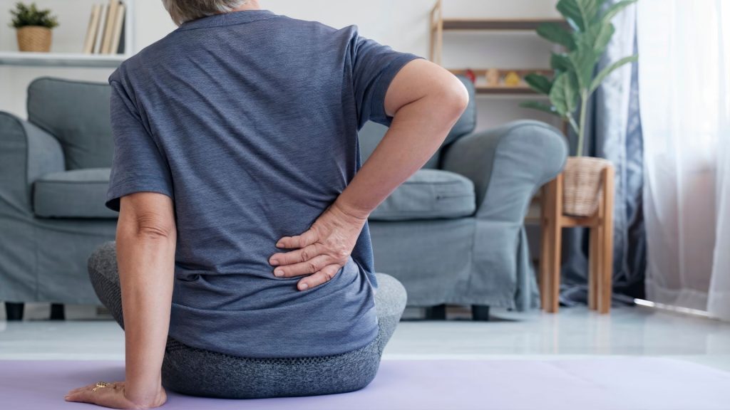 An elderly woman sitting on a yoga mat, holding her lower back in discomfort.