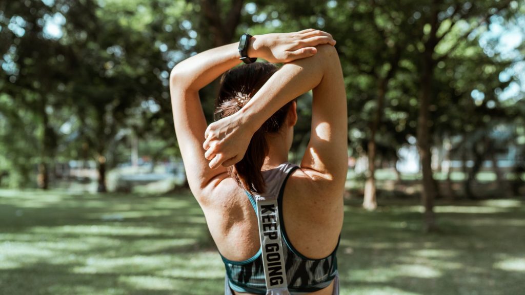 A woman stretching her arms in a park, wearing a sports top with "KEEP GOING" printed on the back.