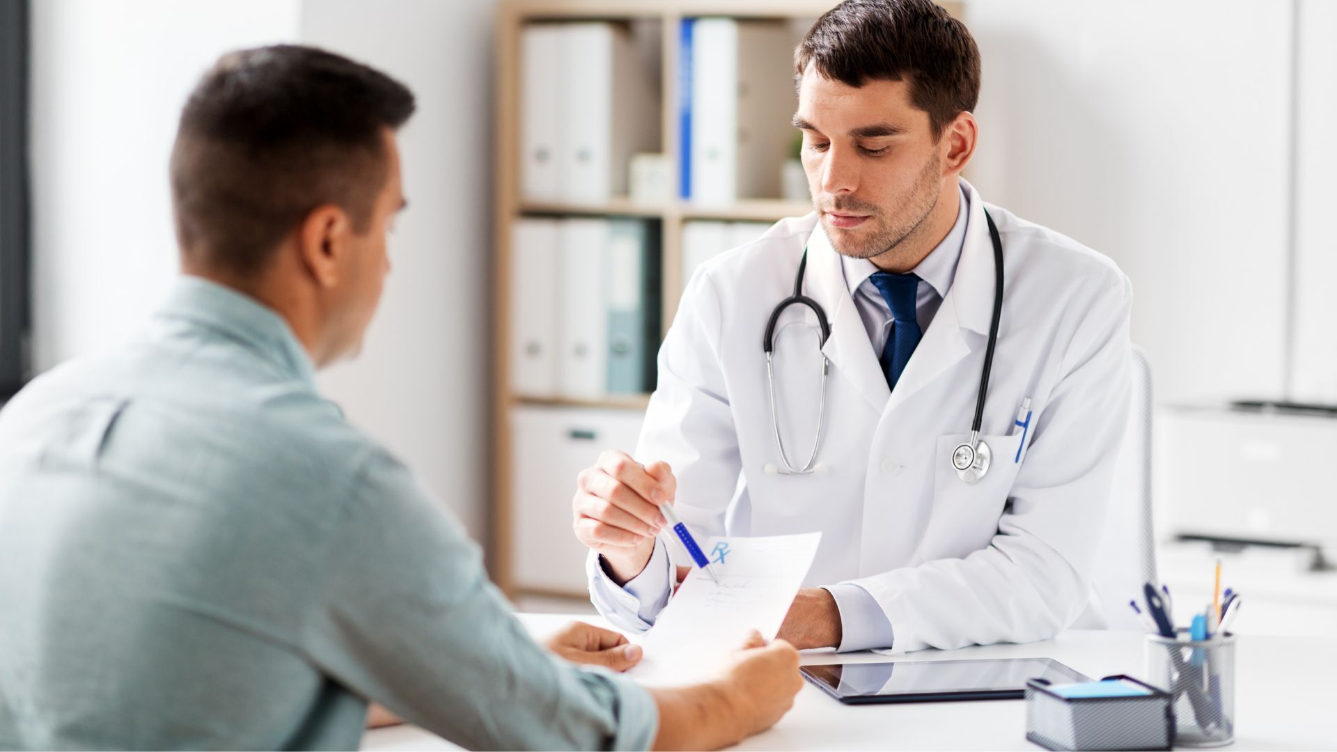 A doctor in a white coat explaining a prescription to a male patient in a medical office.