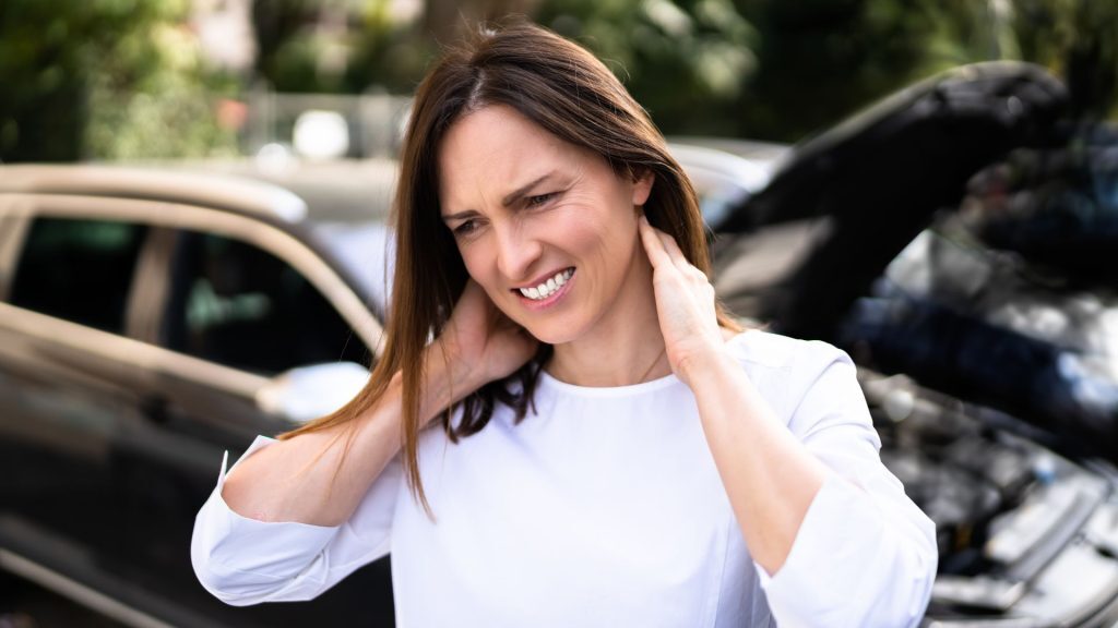 Woman standing near a car accident, holding her neck in pain.