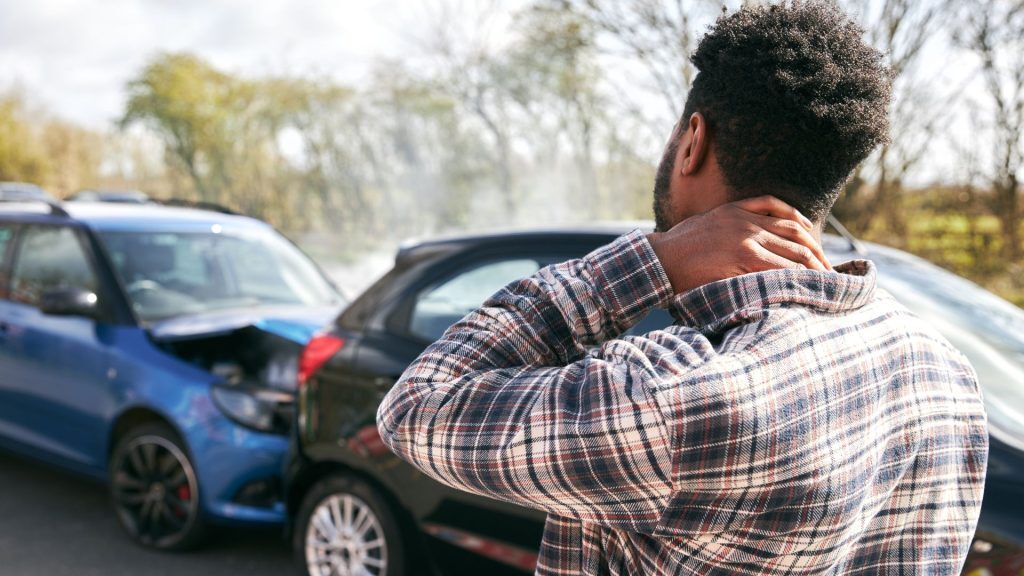 Man holding his neck after a car crash with damaged vehicles in the background.