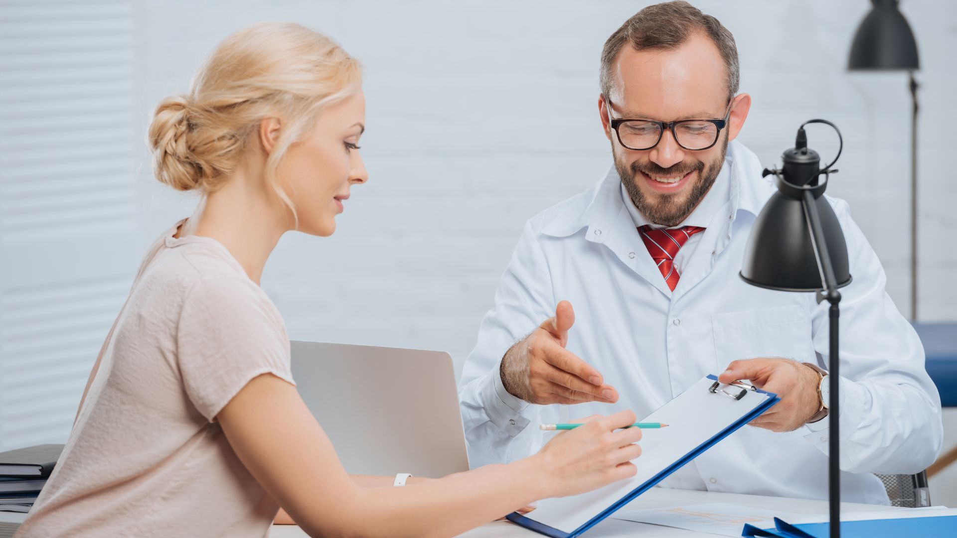 Female patient and smiling male doctor discussing documents during a consultation in a well-lit clinic office, suggesting follow-up care or treatment planning.