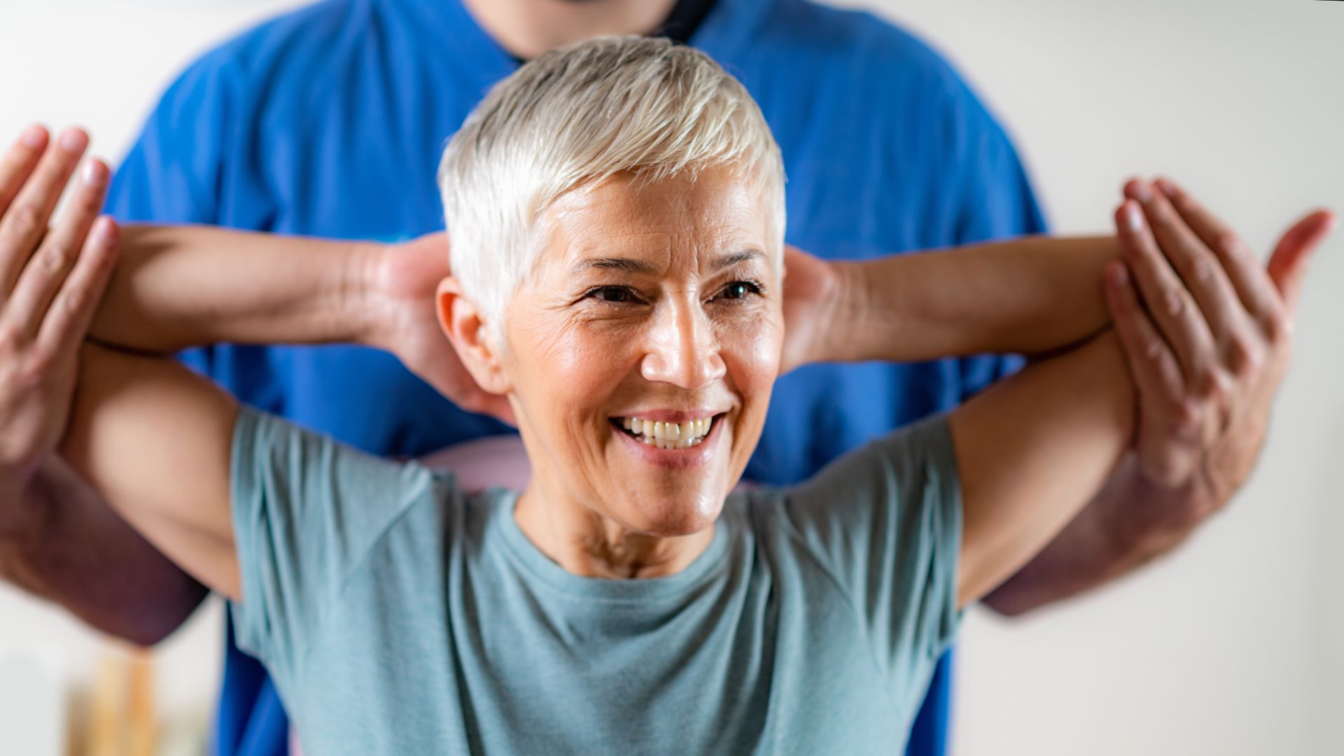 Smiling older woman stretching her arms with assistance from a physical therapist, indicating recovery or guided rehabilitation.