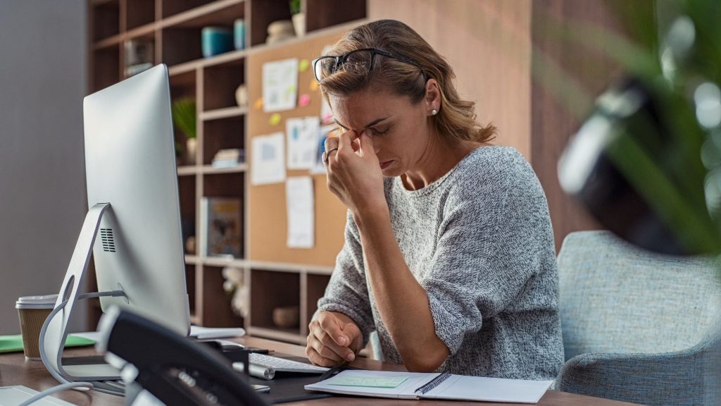 Woman sitting at a desk with her eyes closed, pinching the bridge of her nose, indicating a headache or stress.