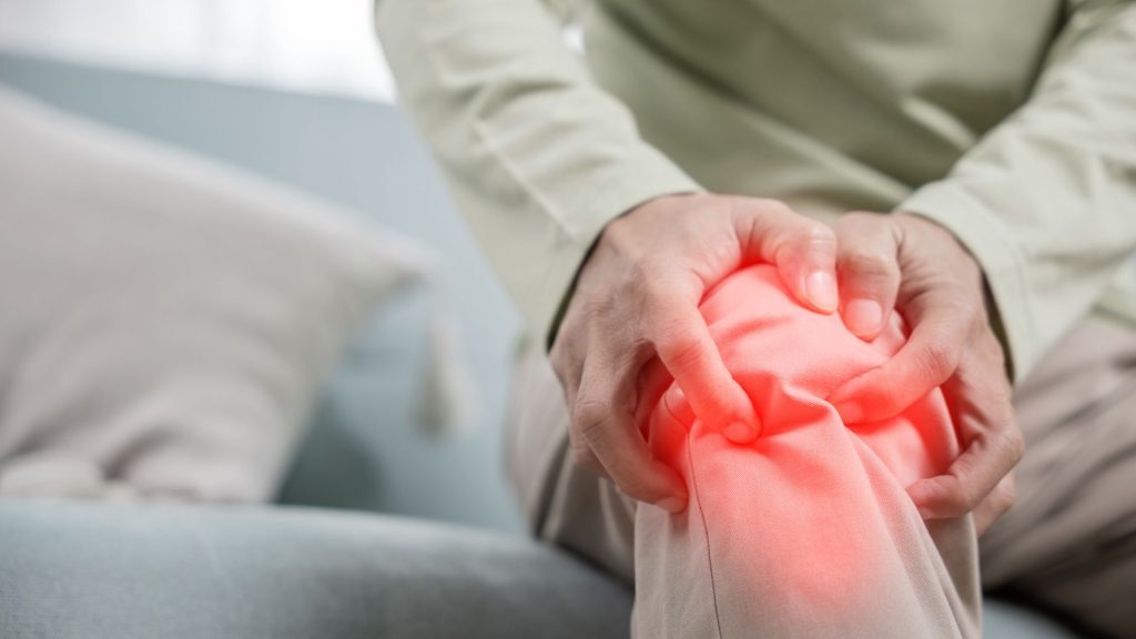 A close-up of an older adult seated on a couch, clutching their knee in discomfort, with a red glow effect highlighting the area to indicate joint pain or stiffness.
