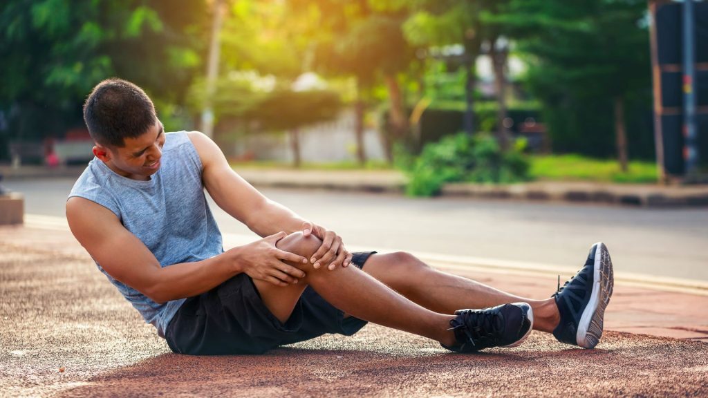 A young man sitting on an outdoor track, wincing in pain as he holds his knee, possibly after a fall or sports-related injury, with a warm sunlight backdrop.