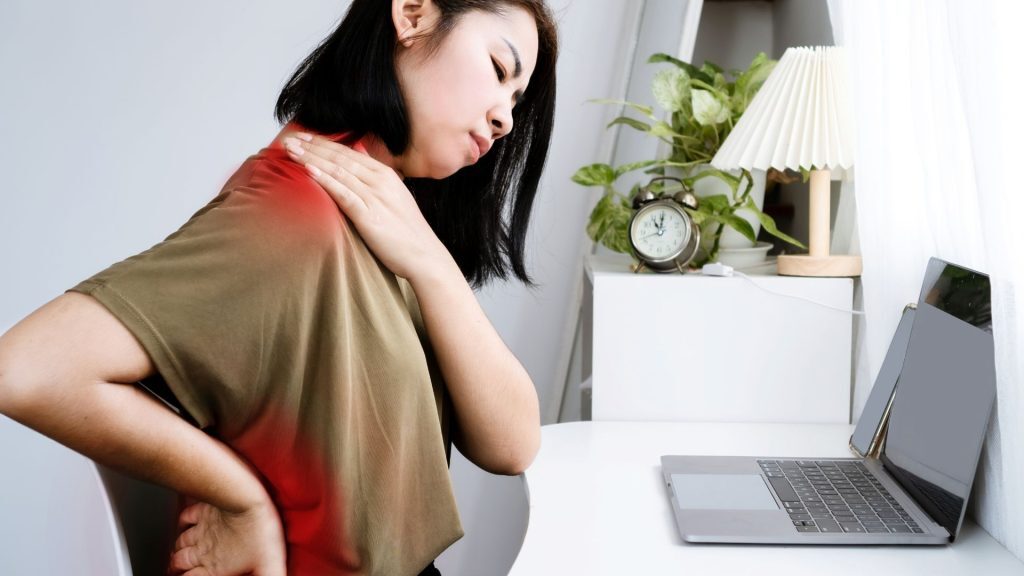 Woman sitting at a desk, holding her neck and back in pain from screen use.
